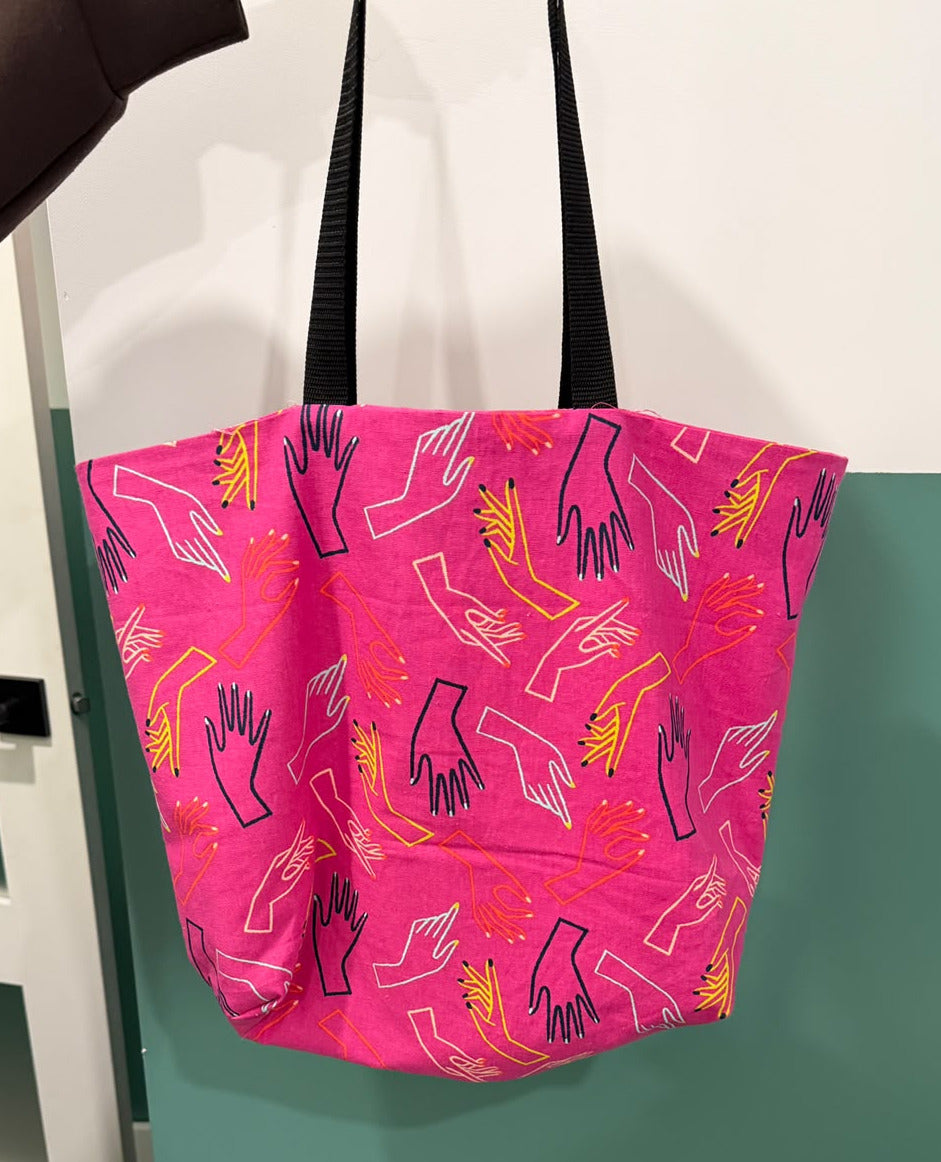 A bright pink tote bag with black handles, featuring a repeating pattern of stylized hand illustrations in black, white, and yellow, hanging against a teal and white wall.